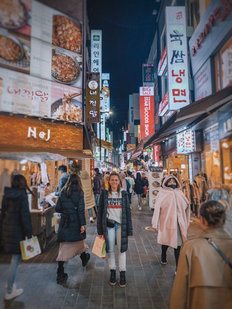 Elyse standing in the middle of a busy street in Seoul, it's night and there are lots of people who travel around her.