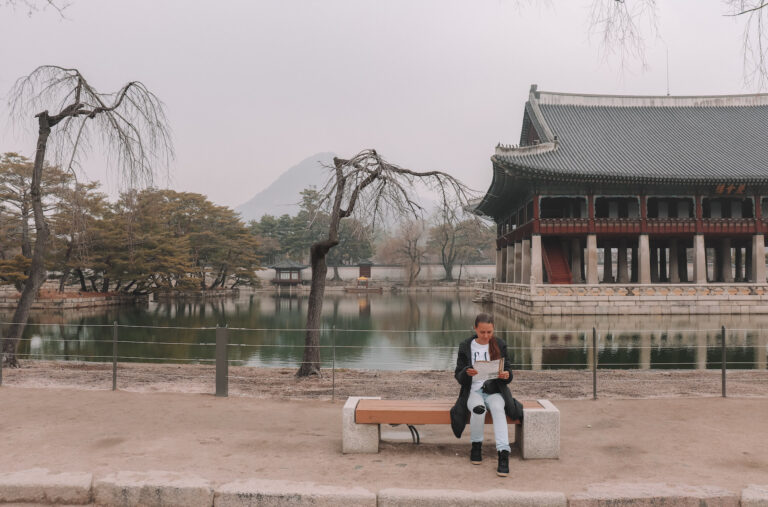 Elyse sitting on a bench reading a map at Gyeongbokgung Palace. An old building on a lake can be seen behind her. She is planning her travel around this Seoul Palace
