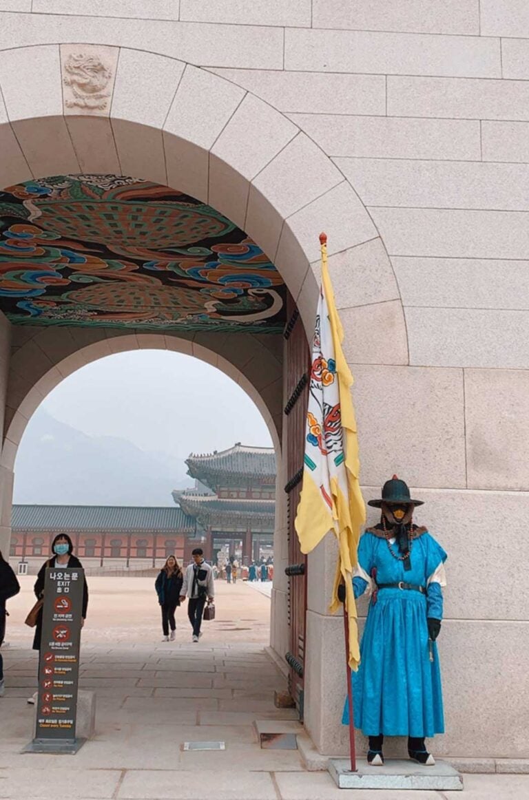 A guard wearing blue and holding a large yellow flag is standing outside Gyeongbokgung Palace