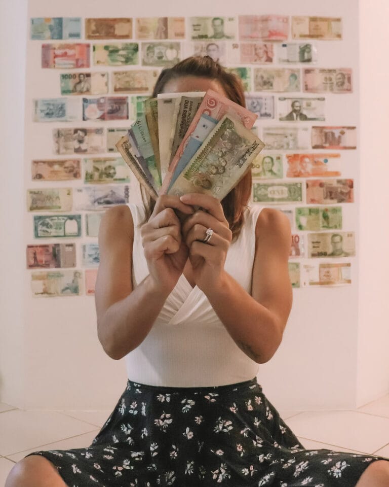 Elyse sitting on the ground holding a fanned out pile of foreign money. On the wall behind her, many different currencies are stuck to the wall