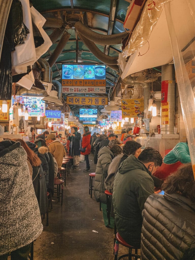 An indoor food market in Seoul, two rows of people are sitting down eating
