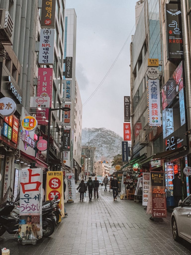 A street in Seoul with a lot of signage and in the distance is a snowy mountain