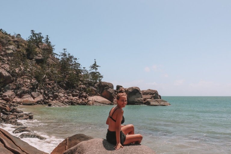 Elyse sitting on a large boulder at the beach on magnetic island. She is smiling at the camera and the ocean behind her looks very clean.