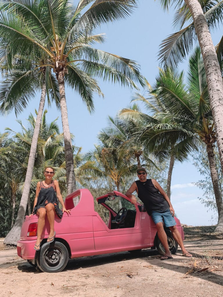 A male and female leaning against a small car that has no roof and is hot pink in color. They’re surrounded by palm trees on Magnetic island.