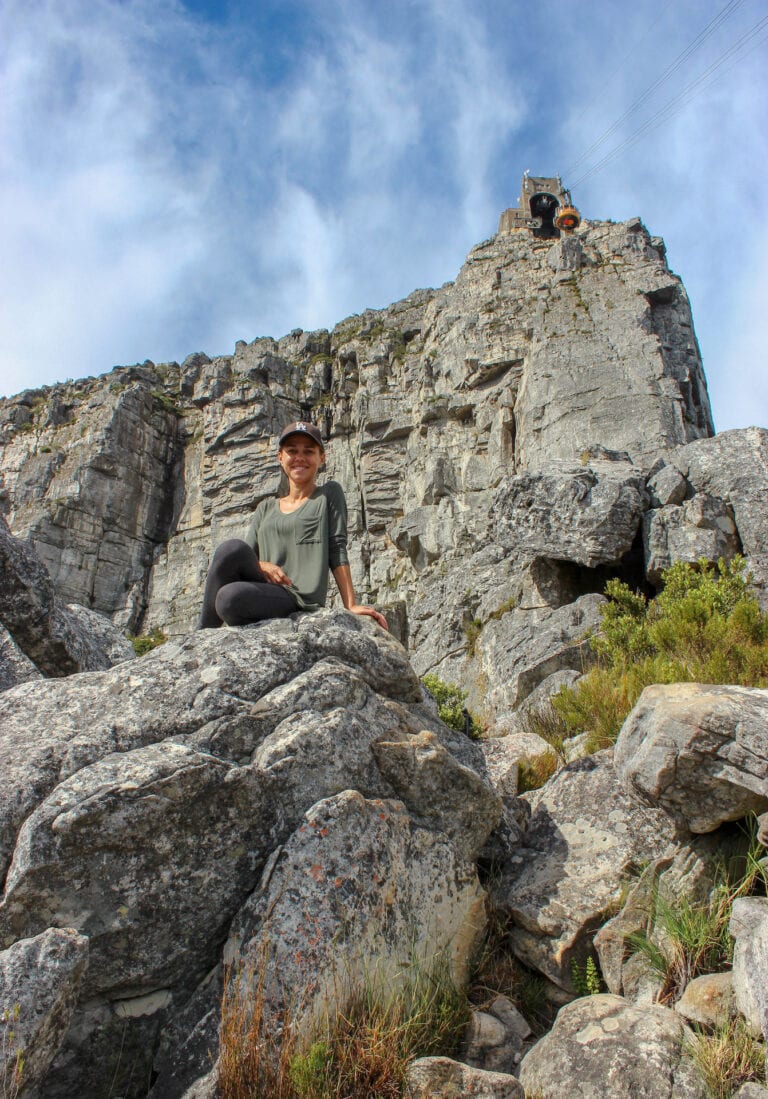 Image looking upwards and Elyse sitting on the edge of a large rock smiling. This was during a hike up Table Mountain and one of my best travel experiences