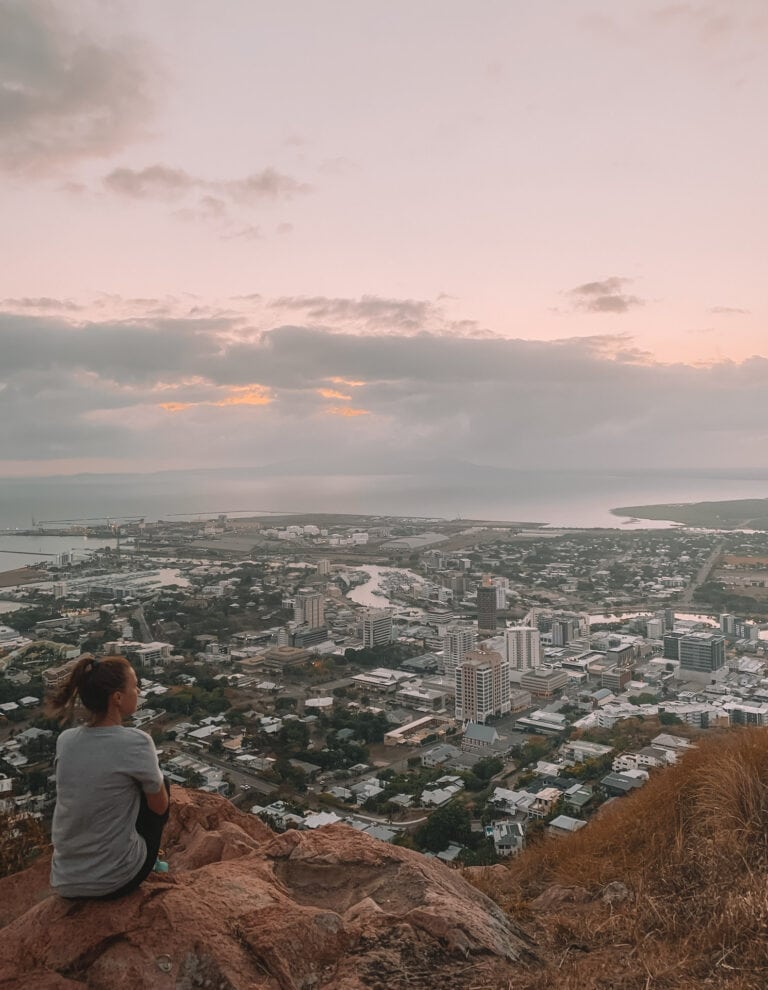 Elyse sitting on the edge of the rocks at the summit of castle hill, looking out at the sunrise over the city of Townsville.