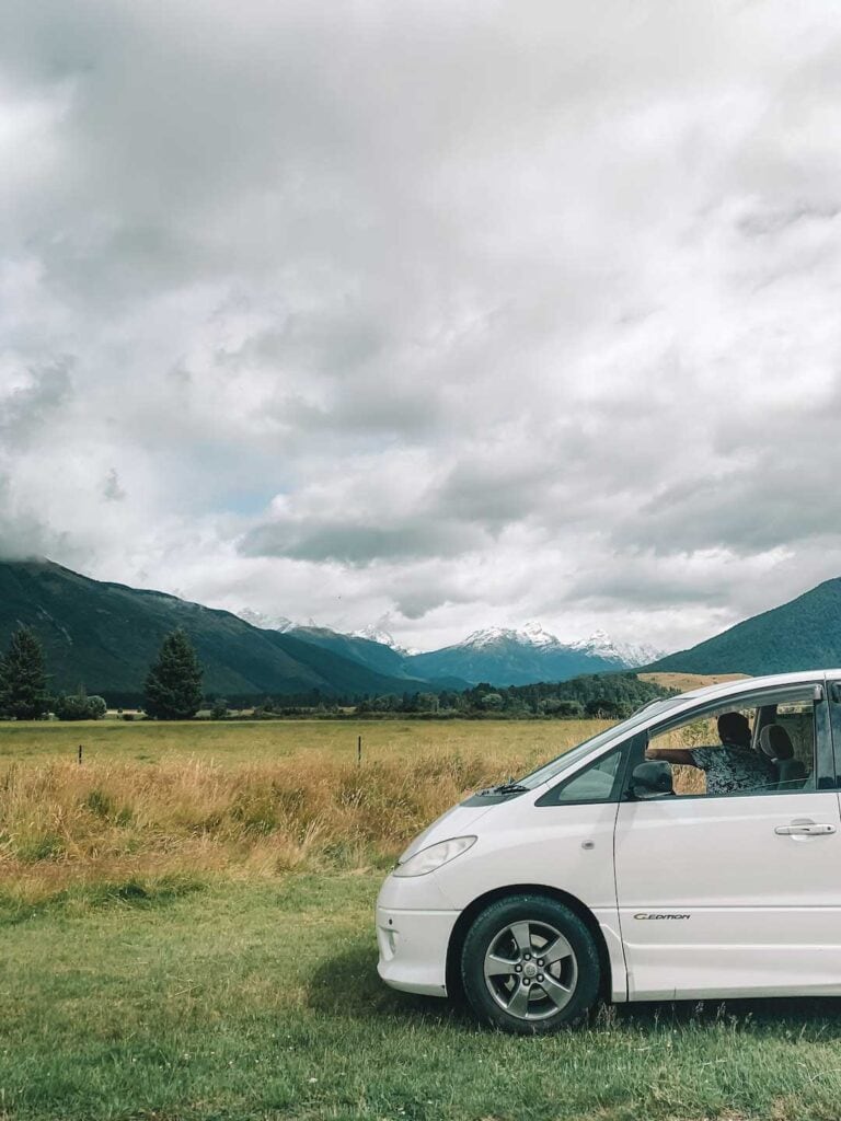 A white Jucy van parked in the wilderness in New Zealand. In the distance snowy mountains can be seen.