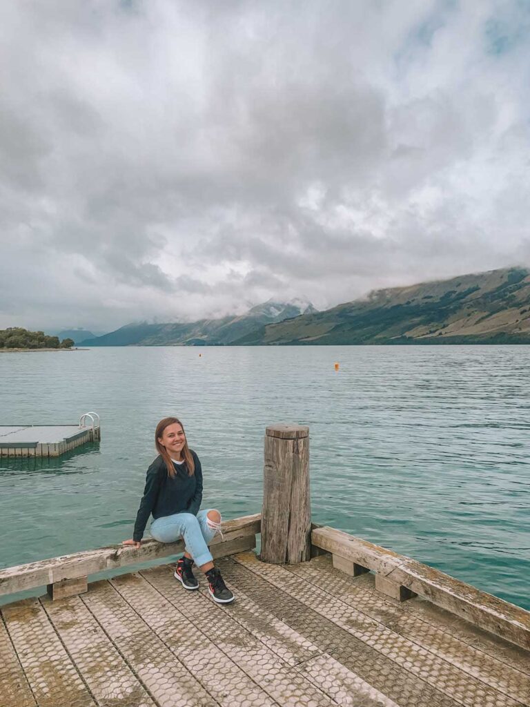 Elyse sitting on the wharf in Glenorchy. It's a cloudy day and there's snowy mountains in the distance