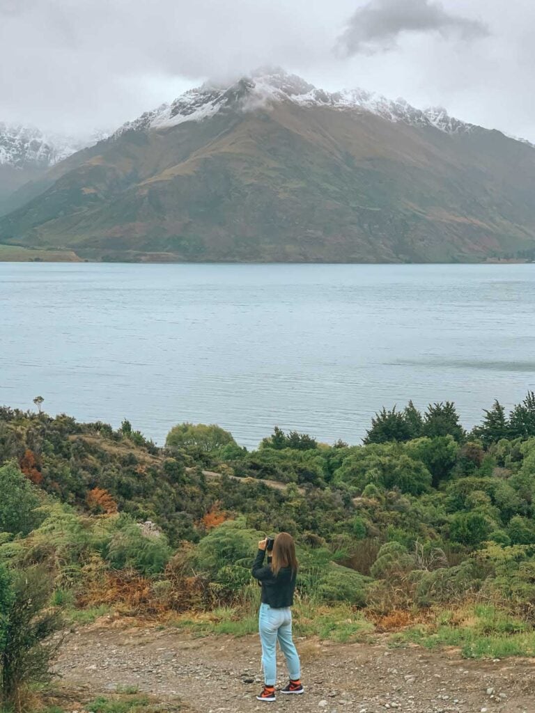 Elyse standing on the lakes edge in Queenstown new Zealand. Taking photos of the snow covered mountains