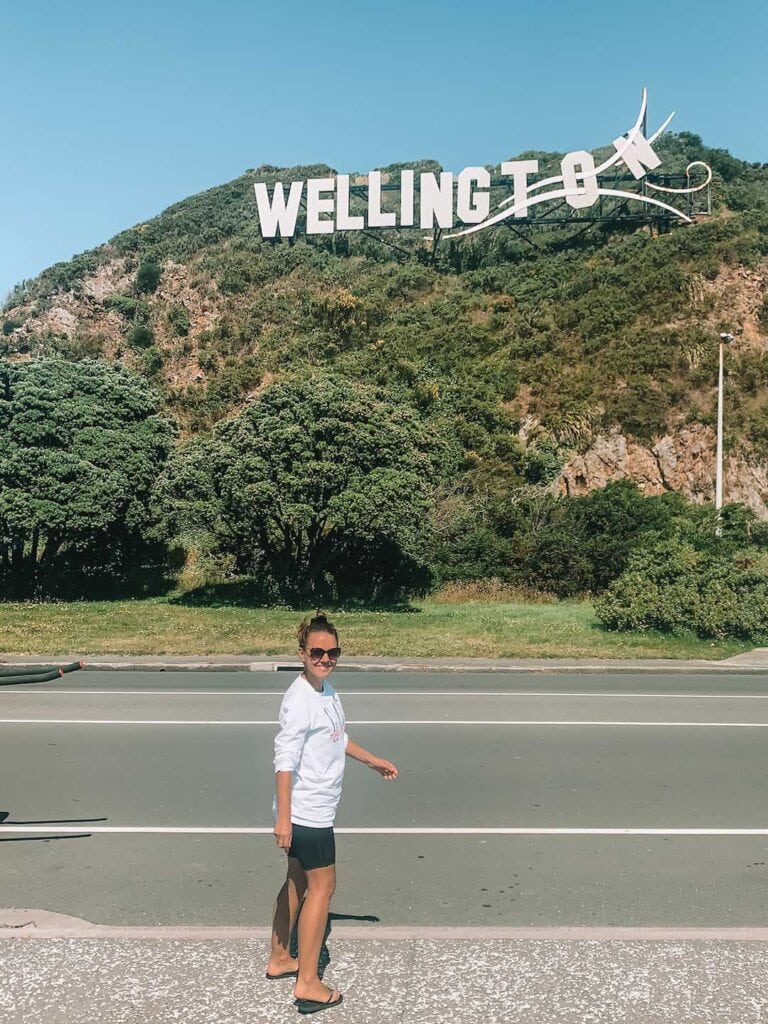 Elyse standing on the side of the road and behind her is green hills and the windy Wellington sign.