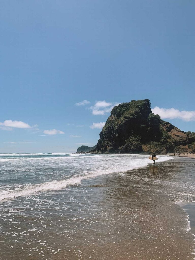 A surfer walking out of the ocean at Piha beach, with Lions Rock in the background. This is one of the activities recommended during a Piha day trip to Auckland
