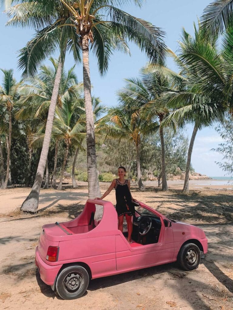 Elyse standing on the front seat of a small topless car, the car is hot pink in color and the area has many palm trees.