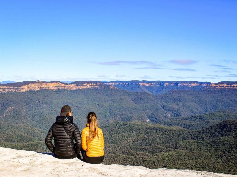 two people sitting on the edge of the cliff looking at the view of the blue mountains in new south whales