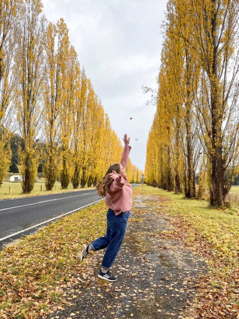 A female on the side of the road jumping and surrounded by yellow autumn leaves. one of the Unique things to do in Melbourne