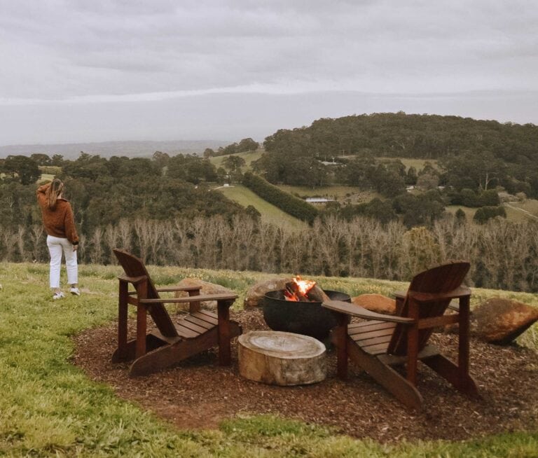 An outdoor setting in the Dandenong Ranges, two wooden chairs facing a fire and over looking rolling green hills on a cloudy day. A female is standing near the seats looking at the view