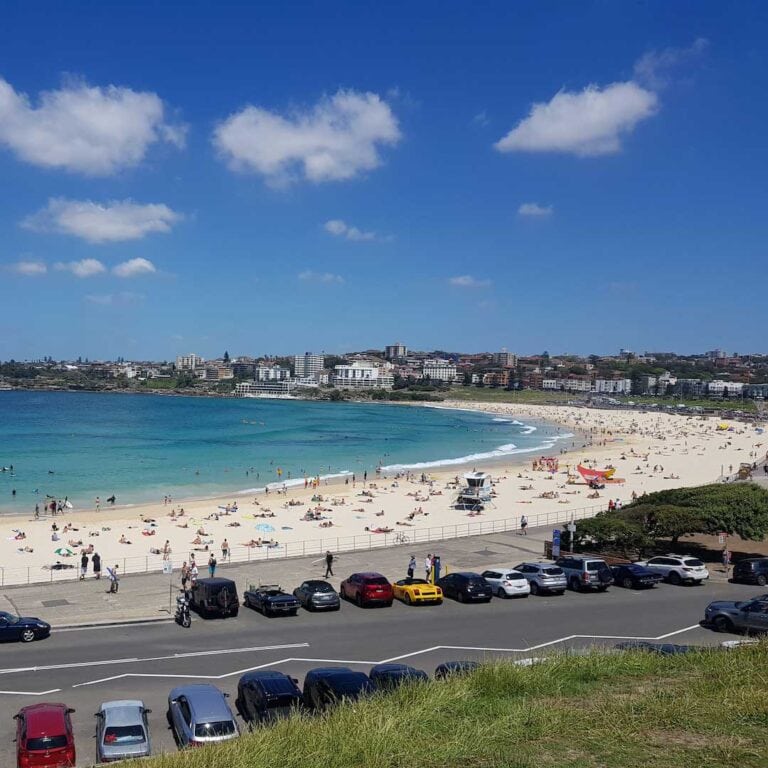 A view of Bondi beach, the beach looks very crowded and there are many cars parked around the beach
