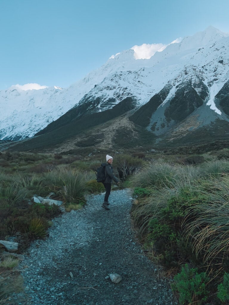 Elyse walking along dirt path in New Zealand. Large snowy mountains in the distance. Packing for a day hike in NZ