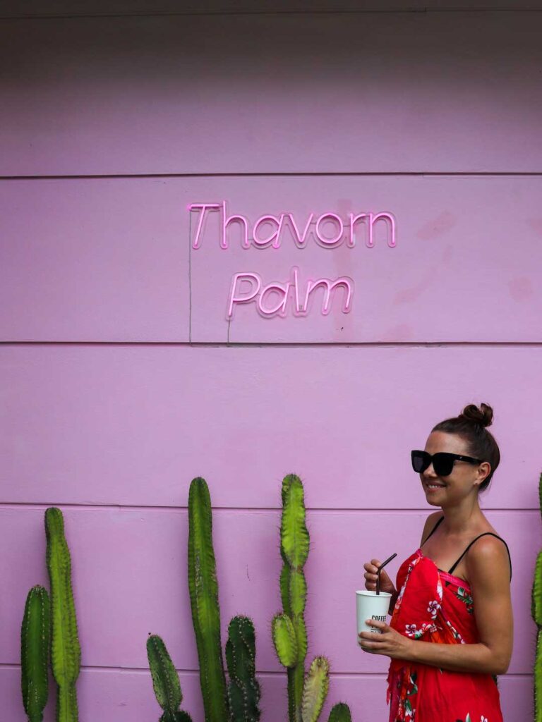 Elyse standing against a pink while drinking an iced coffee at her beachfront accommodation in Karon