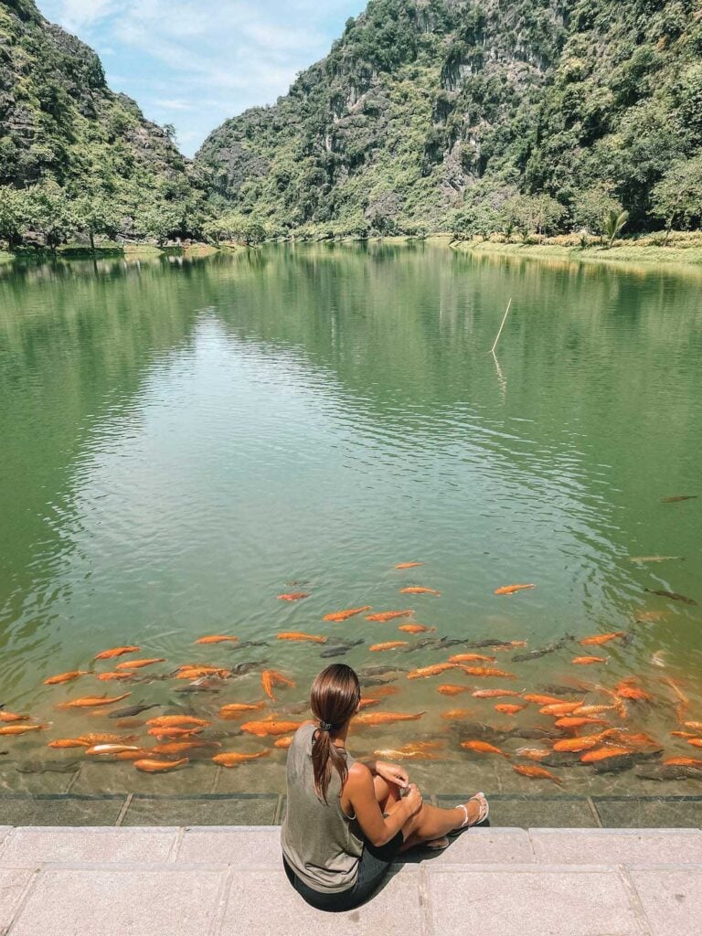 elyse sitting on the edge of a large lake in Am Tien Cave complex in Ninh Binh Vietnam. There are also heaps of gold fish swimming in front of her