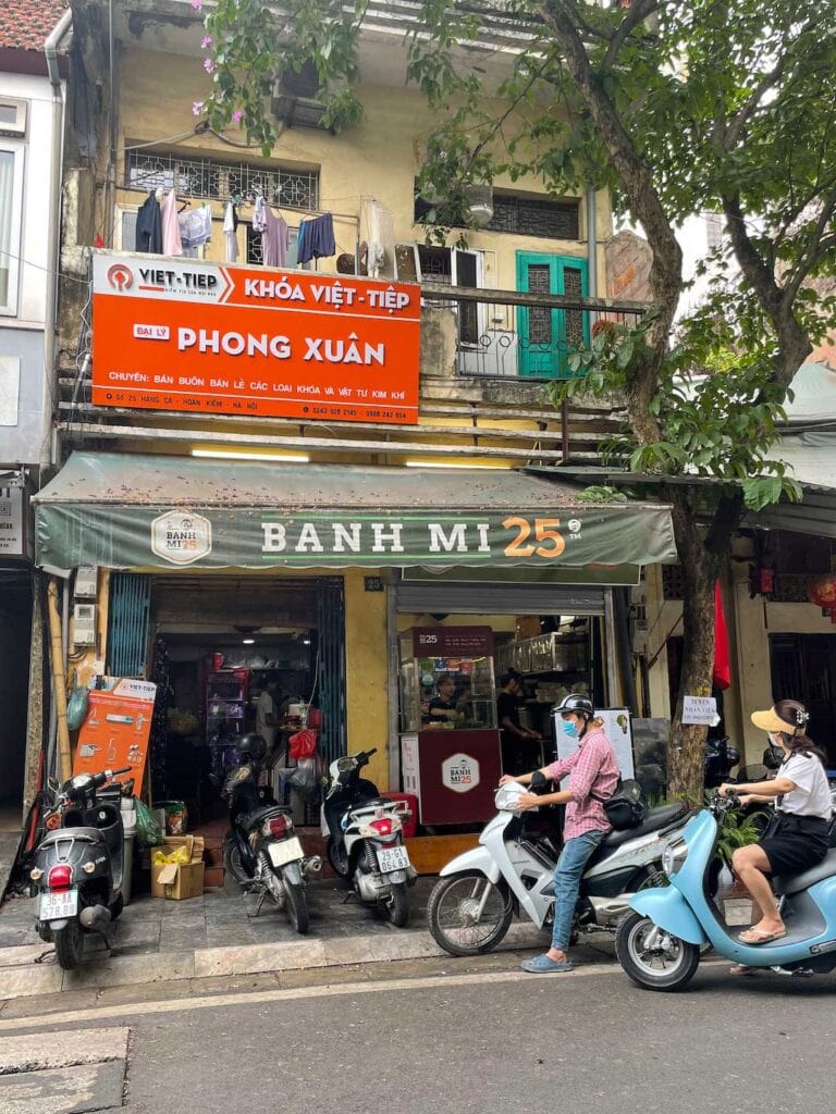 The front of a Banh Mi restaurant in Hanoi, there are 5 motorbikes parked out the front and on the balcony above the restaurant there us washing drying