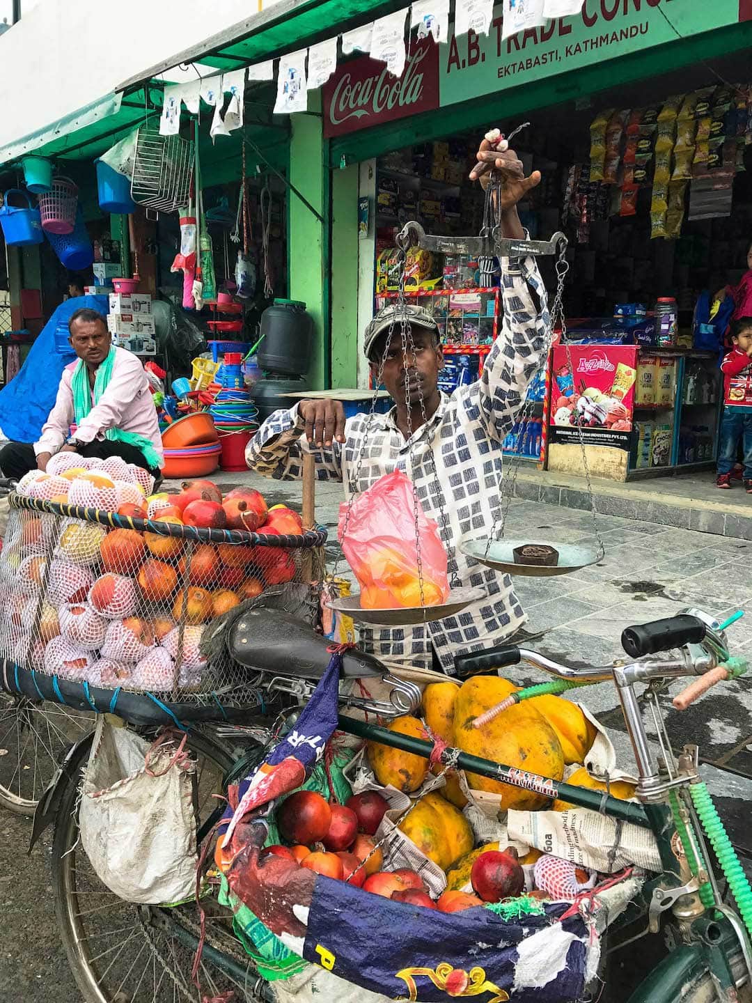Fruit salesmen in Kathmandu. His bicycle is holding the fruit and he is weighing two mangos