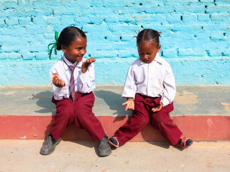 Two young Nepalese Children sitting down in uniform against a blue wall