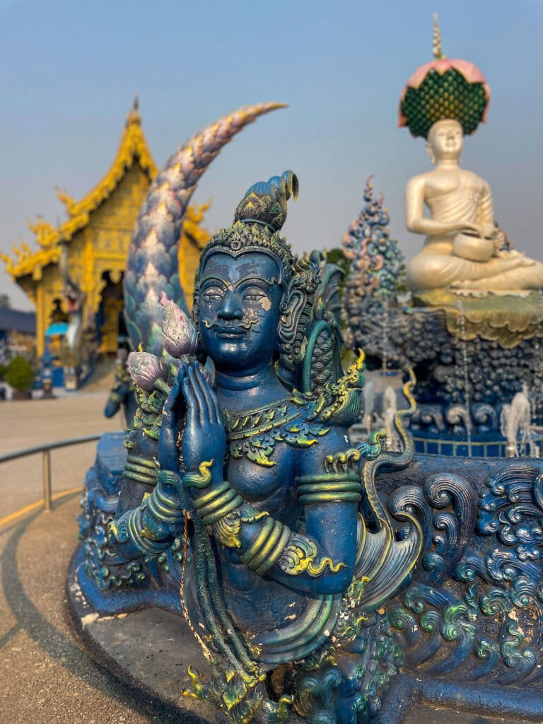 Close-up of blue and gold deity statue at the Blue Temple in Chiang Rai, Thailand, with white Buddha in background.