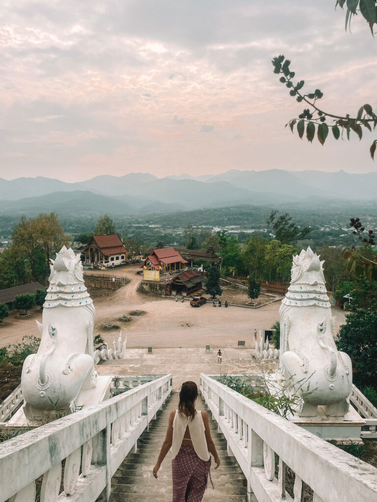Woman walking down white temple stairs in Pai, Northern Thailand, with mountain views at sunset.