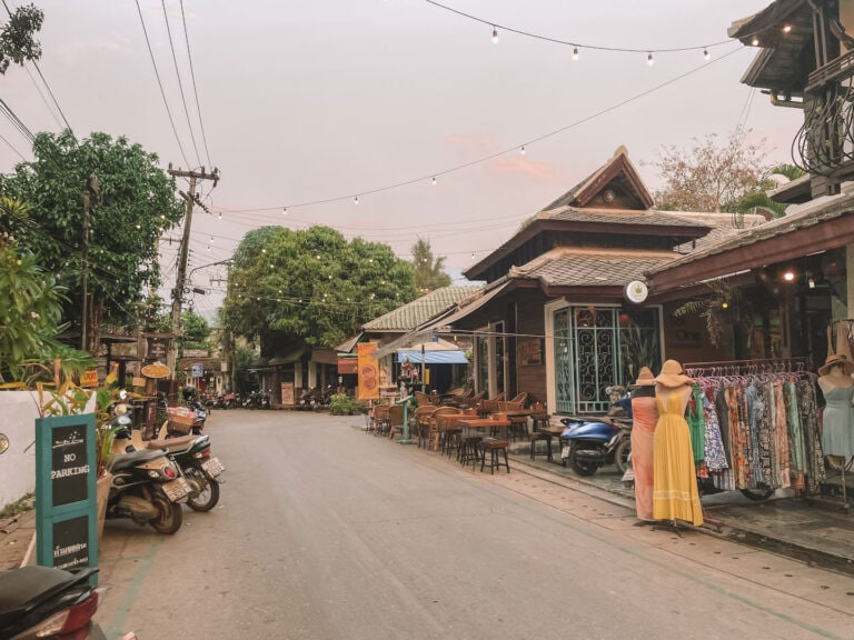 Pai Walking Street in Northern Thailand at sunset, with hanging lights, scooters parked along the road, local shops, and colorful dresses displayed outside.