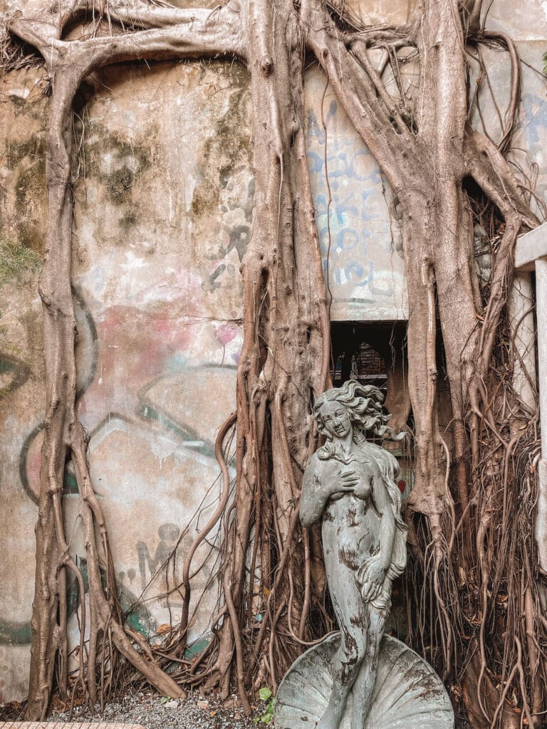 a statue of a woman with the wall behind her covered in tree roots, at Hong-Sieng-Kong-Café