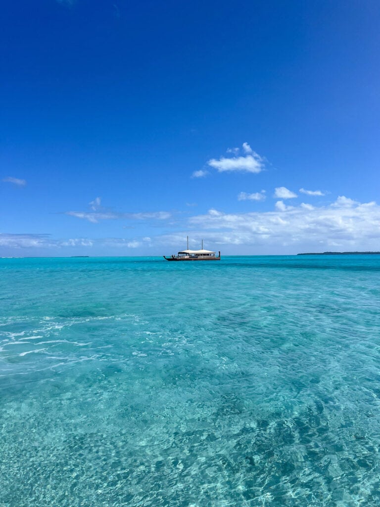 Calm turquoise ocean in Aitutaki Lagoon with a Vaka Cruise under a blue sky.