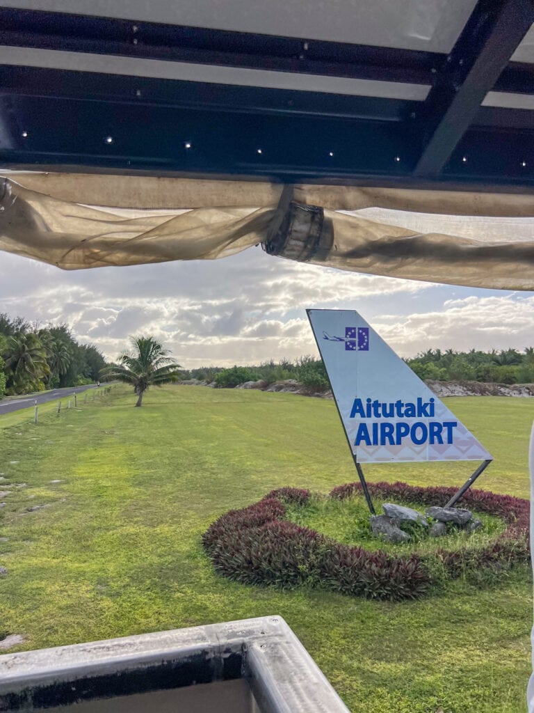 Aitutaki Airport sign with lush green landscape and palm trees in the background.