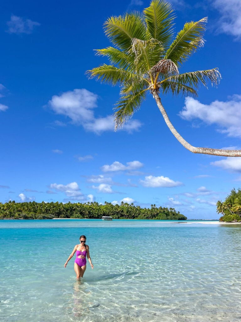 Scenic tropical beach off one foot island in Aitutaki, with palm tree and woman enjoying clear water.