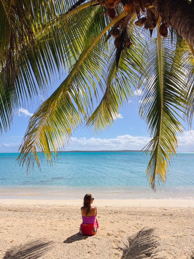 Elyse sitting on sandy beach at Moturakau under palm trees overlooking turquoise ocean during Aitutaki day tour