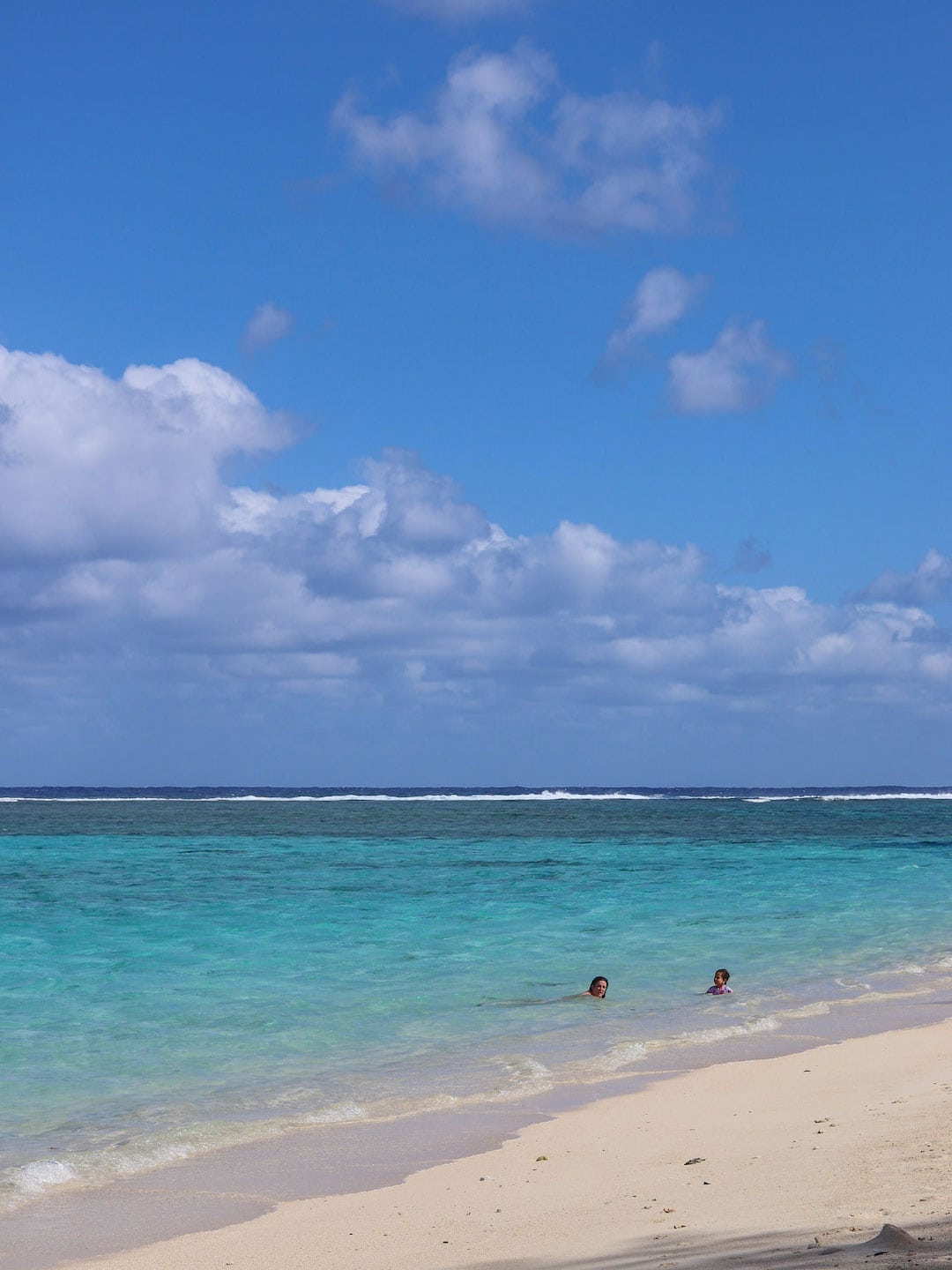 Two people swimming in turquoise ocean waters on a sunny beach in Rarotonga