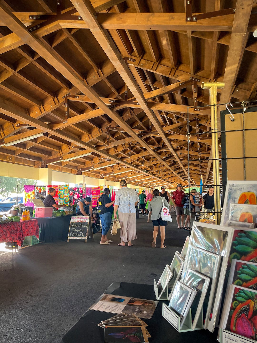People browsing art and crafts at an indoor market under a wooden roof. This is Punanga Nui Market in Rarotonga