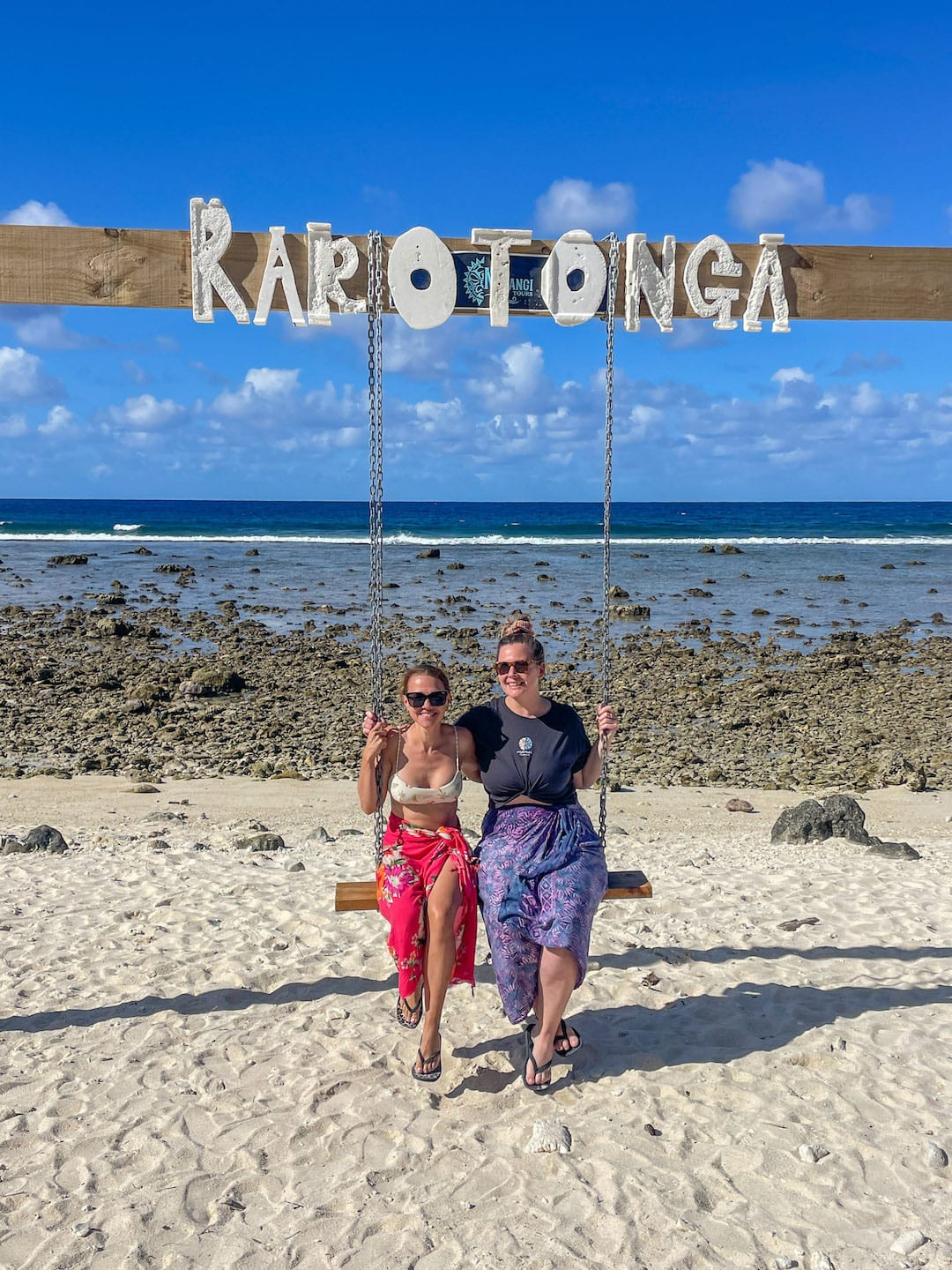 Two women sitting on a beach swing with Rarotonga sign overhead.