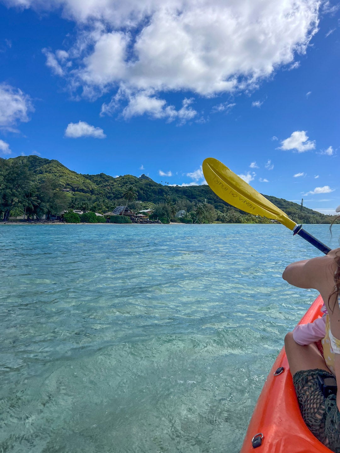 kayaking in the clear tropical waters of Muri Lagoon, with lush green hills and blue sky.